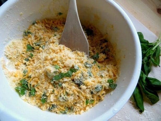  A white bowl with parmesan, cheddar, chopped basil, and other ingredients inside. There is a wooden spoon in the mixture, and a sprig a basil sits in the background.
