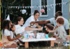 Give Your Outdoor Space a Family-Friendly Facelift Two adults and two children are sitting round a low table in the garden. There is bunting in the background and a variety of food, drinks, plates, and cups on the tabletop.