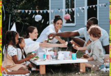 Give Your Outdoor Space a Family-Friendly Facelift Two adults and two children are sitting round a low table in the garden. There is bunting in the background and a variety of food, drinks, plates, and cups on the tabletop.