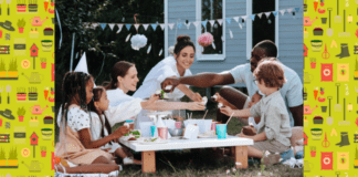 Give Your Outdoor Space a Family-Friendly Facelift Two adults and two children are sitting round a low table in the garden. There is bunting in the background and a variety of food, drinks, plates, and cups on the tabletop.