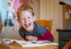 Do You Need a Dedicated Room to Homeschool? A laughing boy looks at the camera while leaning on a book on a table.