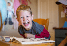Do You Need a Dedicated Room to Homeschool? A laughing boy looks at the camera while leaning on a book on a table.