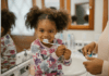 Oral Care Habits to Teach Your Child Early On A smiling child sit on the bathroom counter and brushes her teeth while learning oral care habits.
