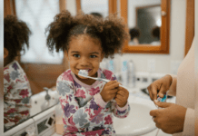 Oral Care Habits to Teach Your Child Early On A smiling child sit on the bathroom counter and brushes her teeth while learning oral care habits.