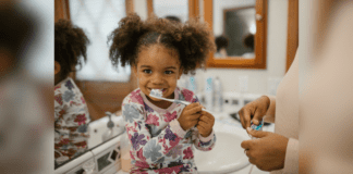 Oral Care Habits to Teach Your Child Early On A smiling child sit on the bathroom counter and brushes her teeth while learning oral care habits.