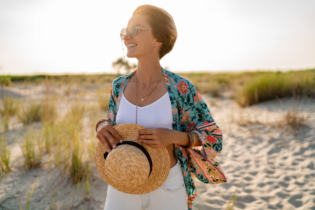 Smiling woman on the beach, wearing white top, jeans and colorful printed tunic boho style chic and straw hat