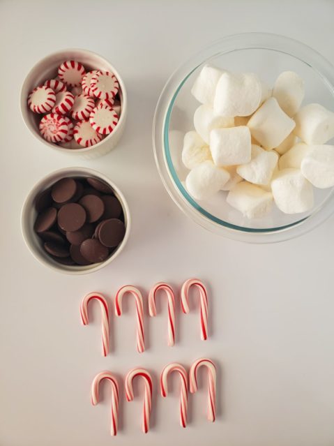 8 mini peppermint candy canes lay on a marble background nest to athree bowls, one with marshmallows, one with peppermints, and one with chocolate.