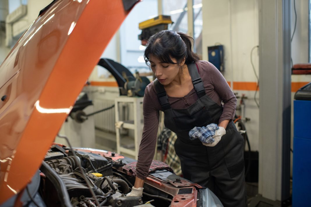 Young female worker of car maintenance service repairing vehicle while bending over open hood with broken engine