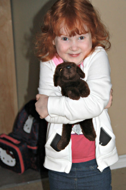Young redheaded girl in jeans, pink t-shirt, and white hoodie, stands holding a stuffed brown dog cuddly toy.